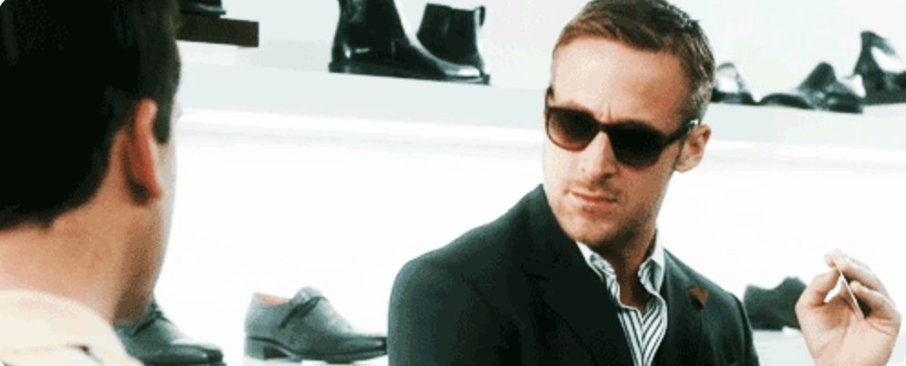 A film still of a man in sunglasses and a dark suit seated across from another man, with shelves of dress shoes on the wall behind them