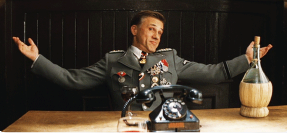 A film still of a uniformed man at a table with a rotary phone and a wine bottle, arms flung wide in an exaggerated shrug